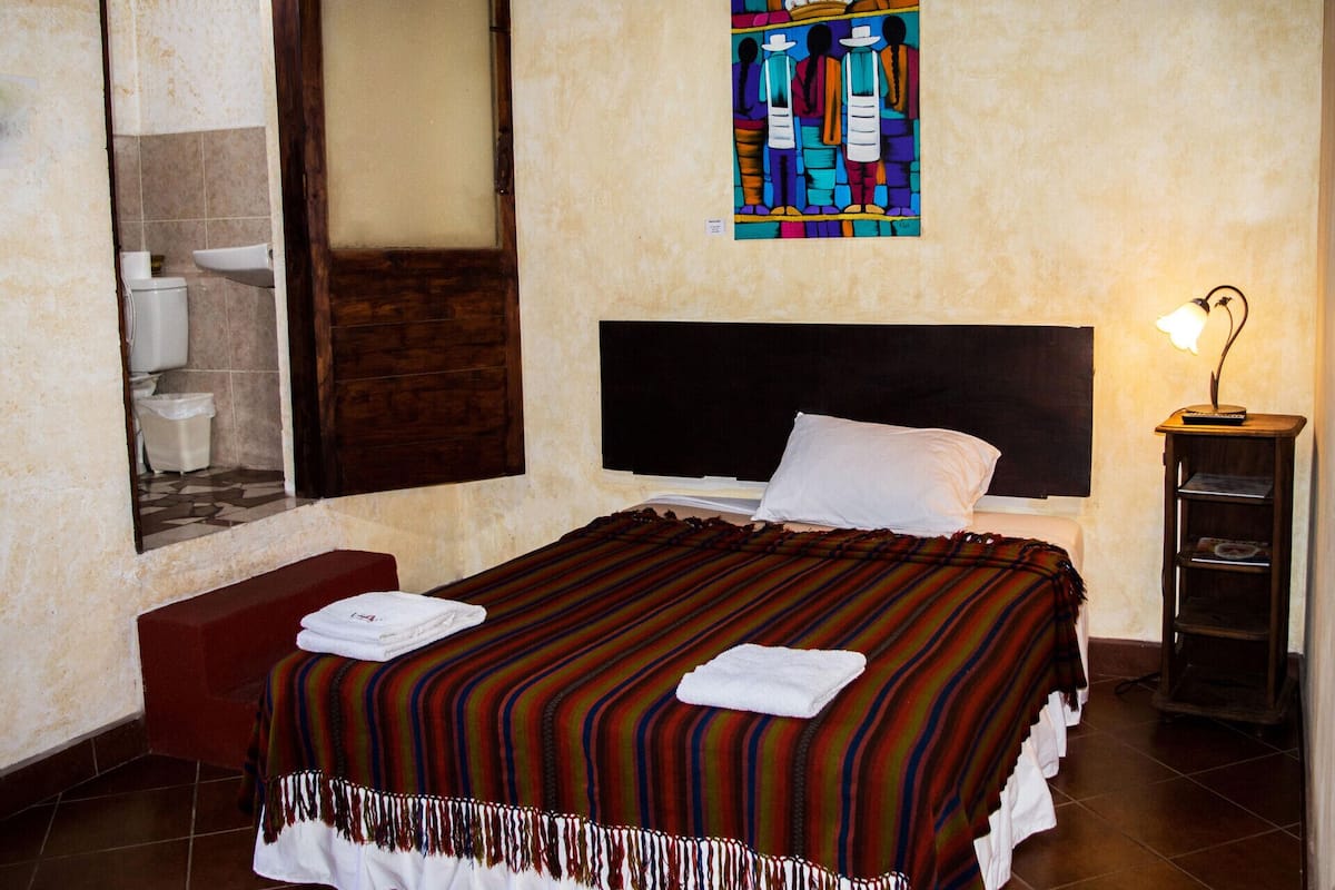 Une chambre d’hôtel avec un lit, une table de chevet, un tableau, une salle de bain visible à travers une porte en verre et une lampe.