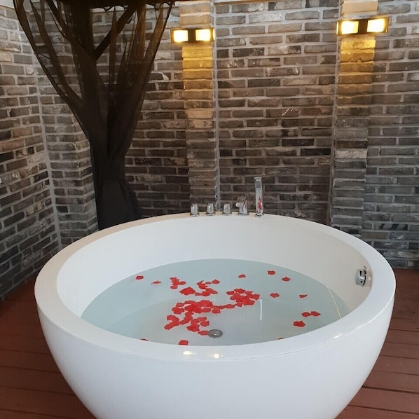 Une grande baignoire blanche remplie d’eau et de pétales de rose, placée devant un mur de briques et éclairée par deux lampes fixées au mur.