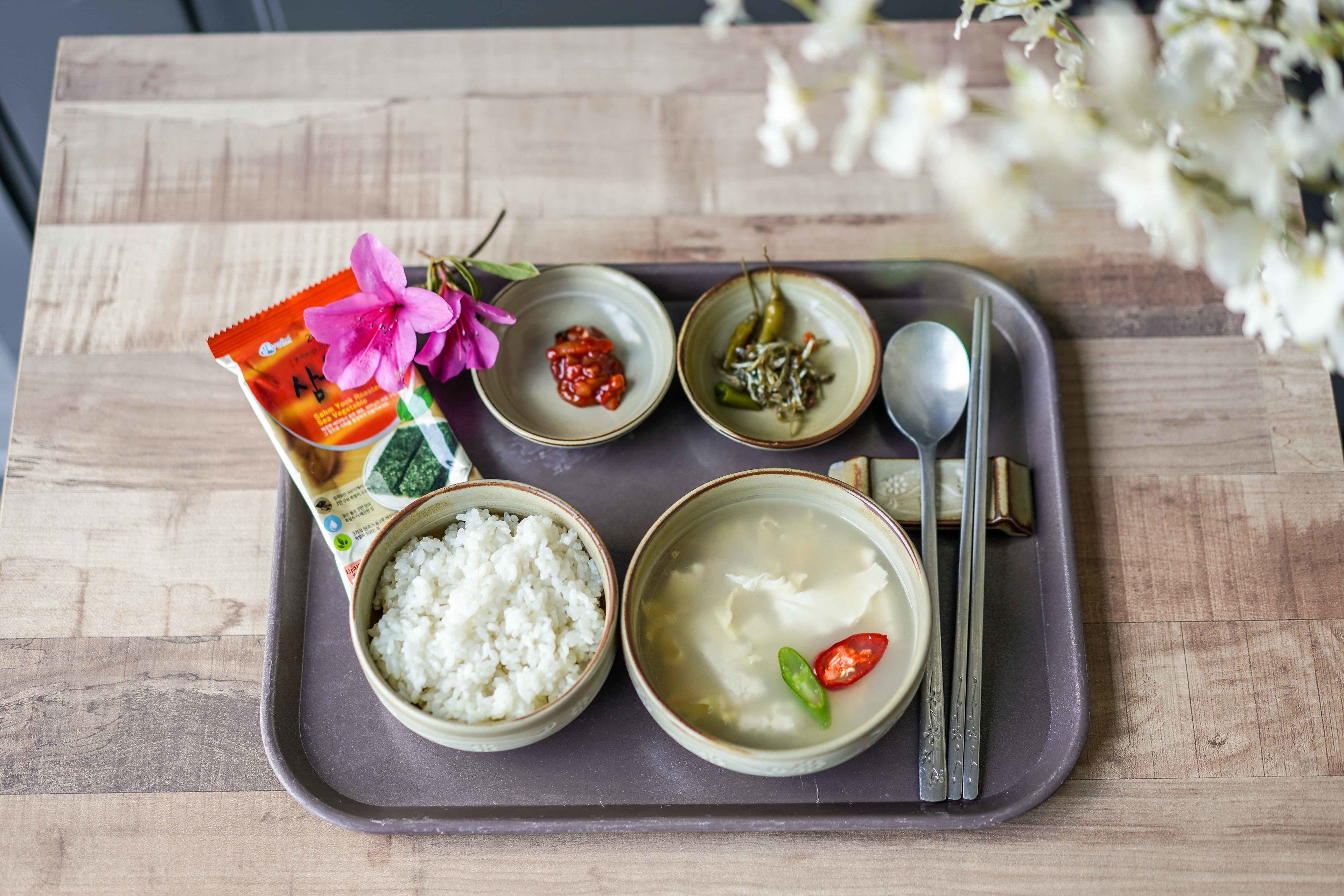 A tray with a package of rice, a bowl of rice, a bowl of soup, a small bowl of chili, and a spoon.