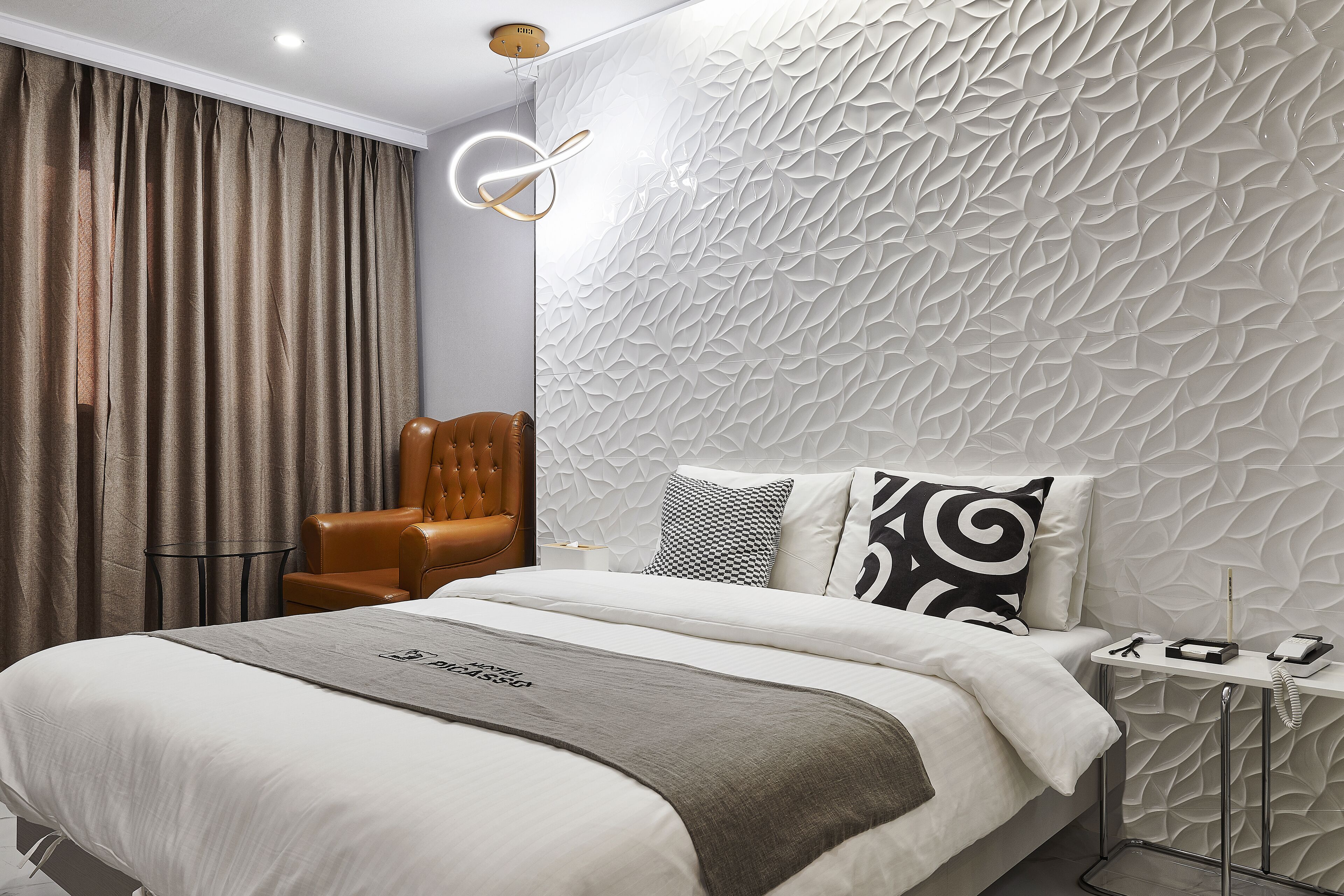 Ein modernes Hotelzimmer mit einem großen Bett, einem Ledersessel, einer strukturierten Wand und einem Nachttisch mit Telefon und Lampe.