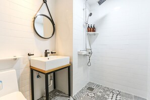 A bathroom with a white sink, mirror, and toilet, a shower with a shelf holding toiletries, and patterned floor tiles. - Picasso Hotel (Donghae)