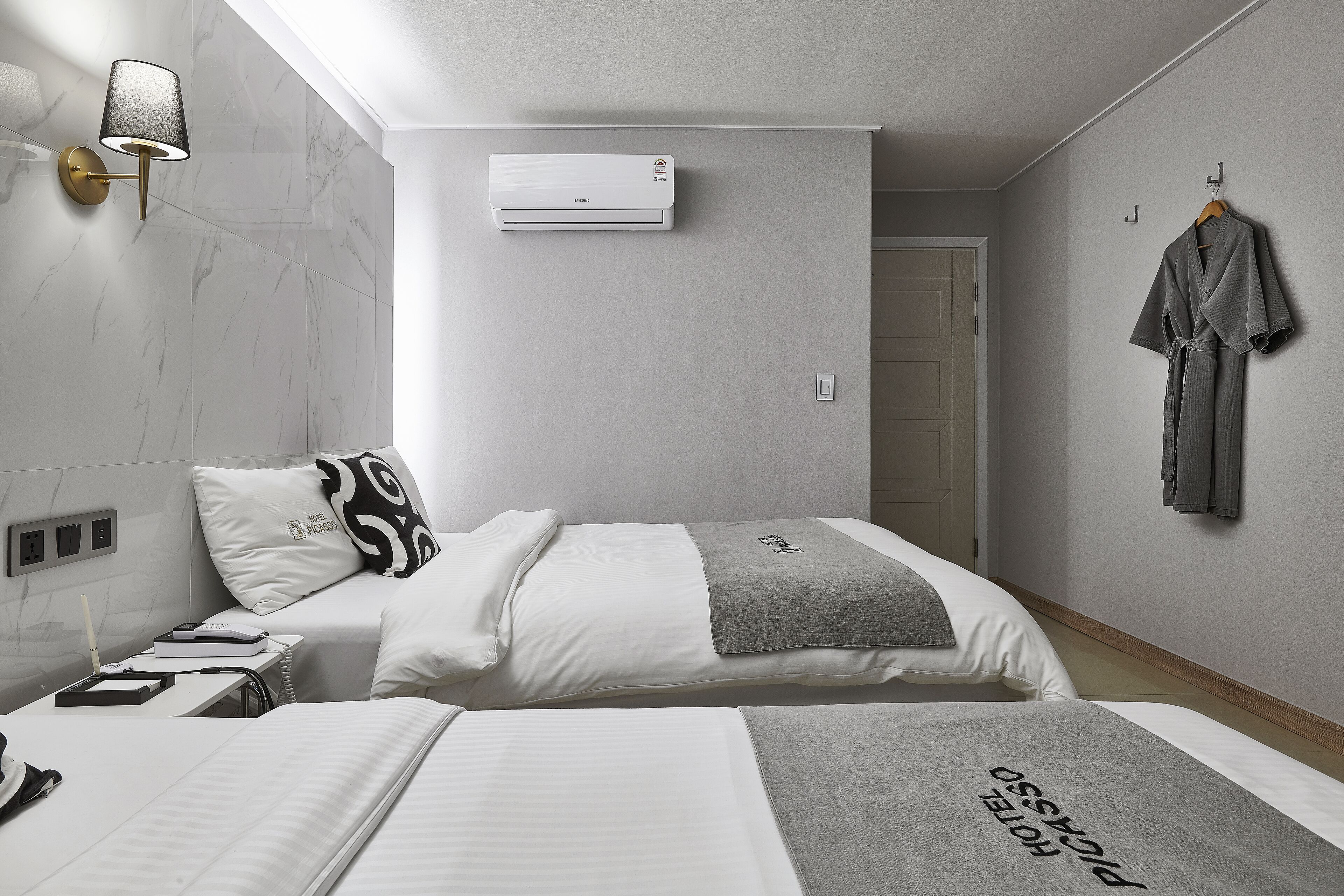 Habitación de hotel con dos camas, una mesita de noche, una lámpara, aire acondicionado y una bata colgada en la pared.