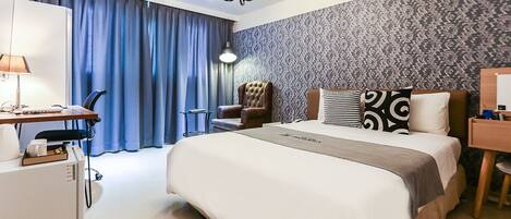 Habitación de hotel con una cama grande, un escritorio con lámpara, una silla y un minibar.
