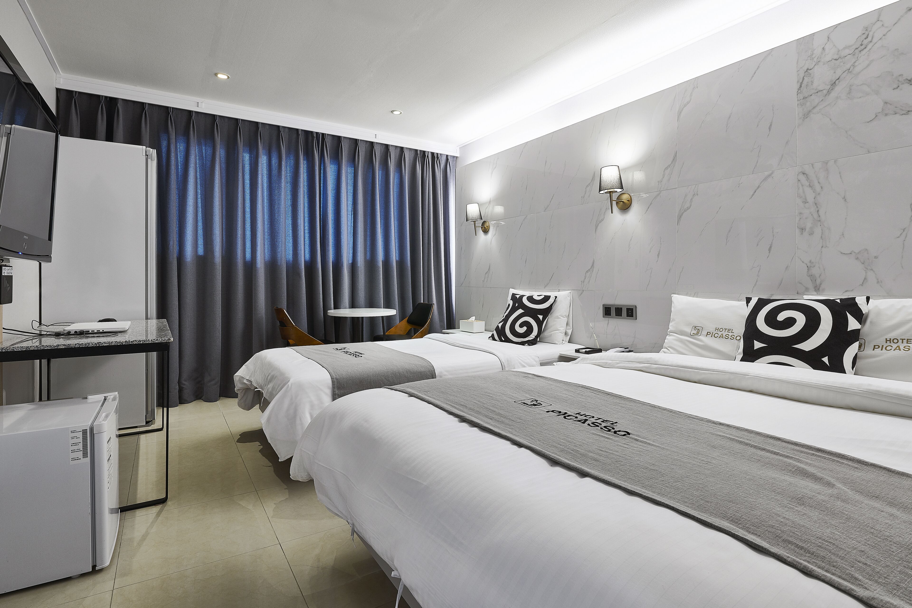 A hotel room with two beds, a desk with a computer, a refrigerator, and a large window with curtains.