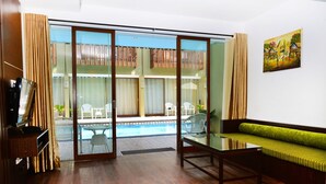 A room with a wooden floor, a glass table, a green cushioned sofa, a television, and a sliding glass door leading to an outdoor area with a pool and chairs. - Devata Suite & Residence (Legian)