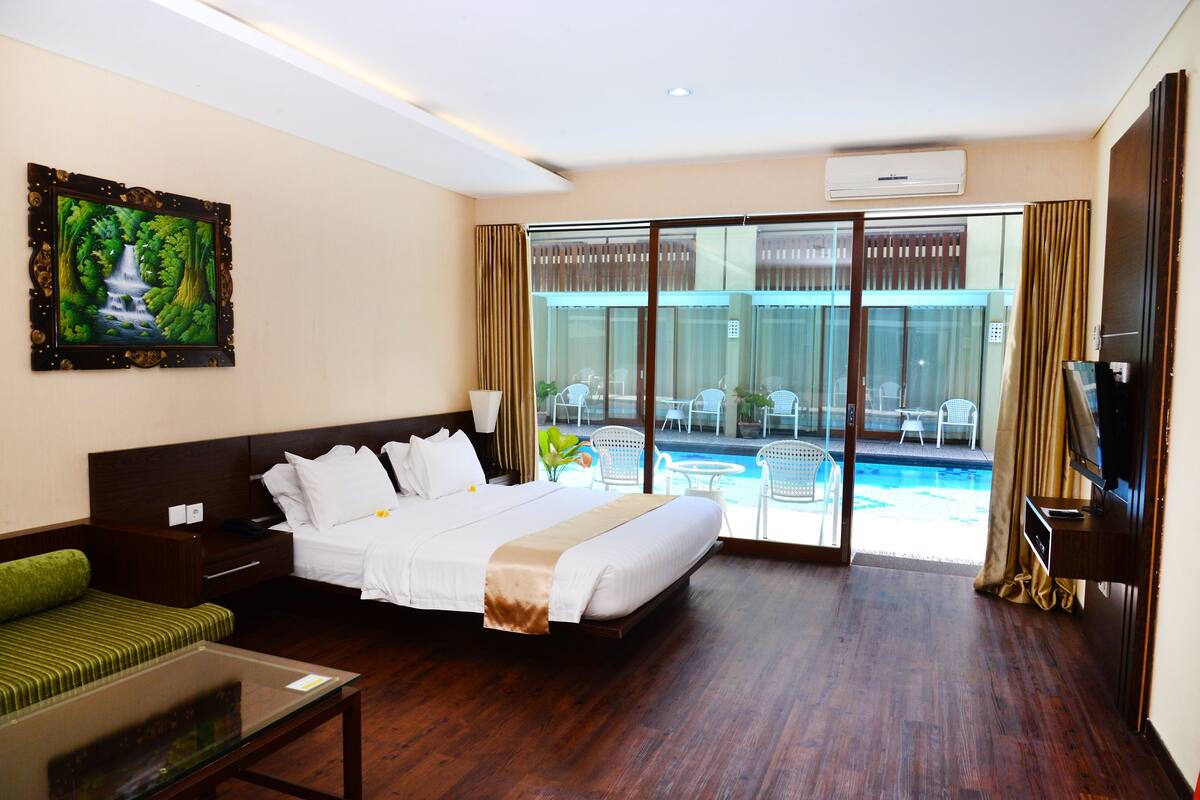 A hotel room with a bed, a glass door leading to a pool area, a TV, and a painting on the wall.