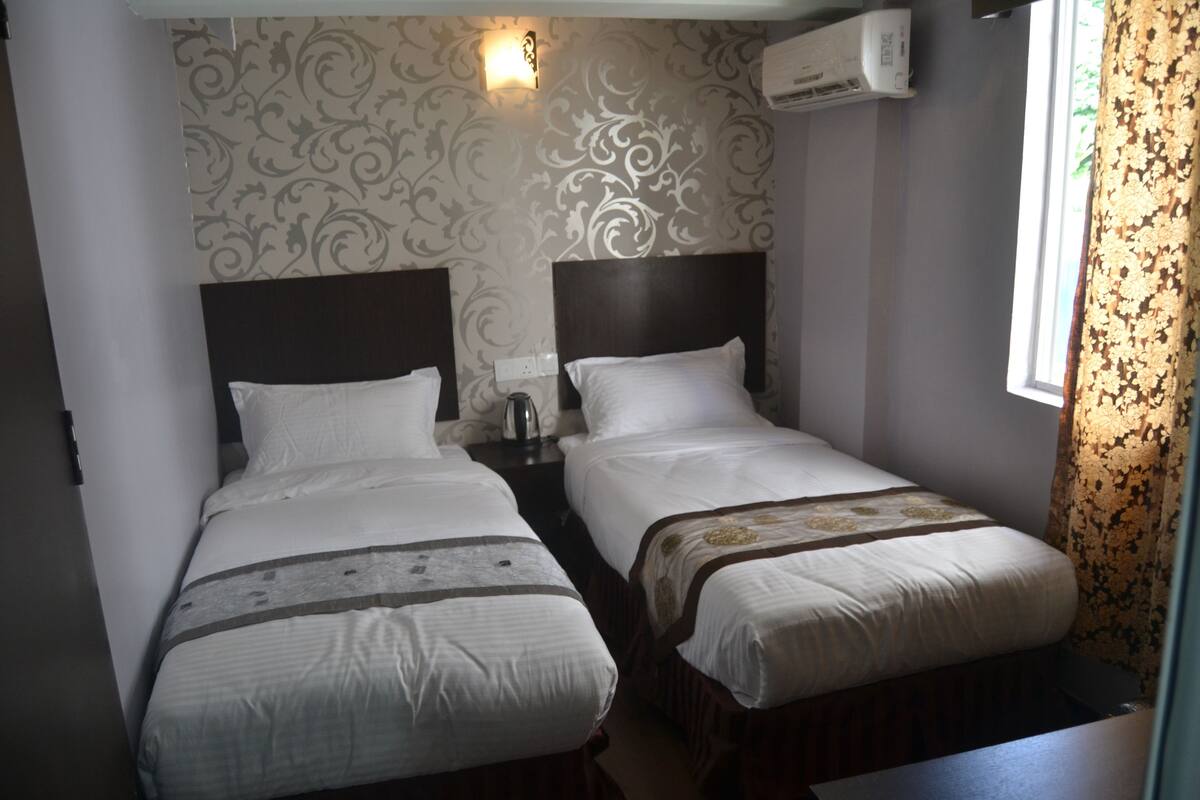 A hotel room with two single beds, a nightstand with a lamp, and a window with floral curtains.