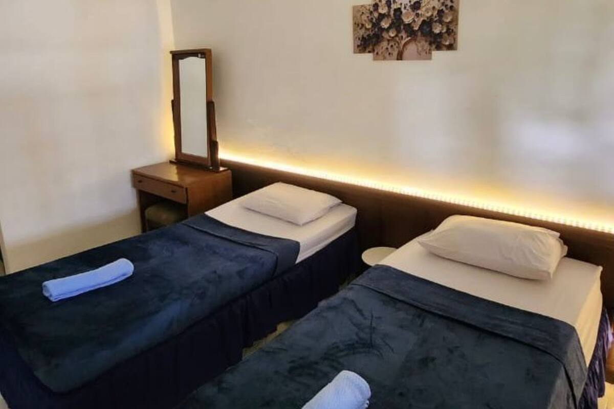 A hotel room with two single beds, each with a dark blue bedspread and white pillows, a small wooden nightstand with a mirror, and a wall-mounted artwork above the beds.