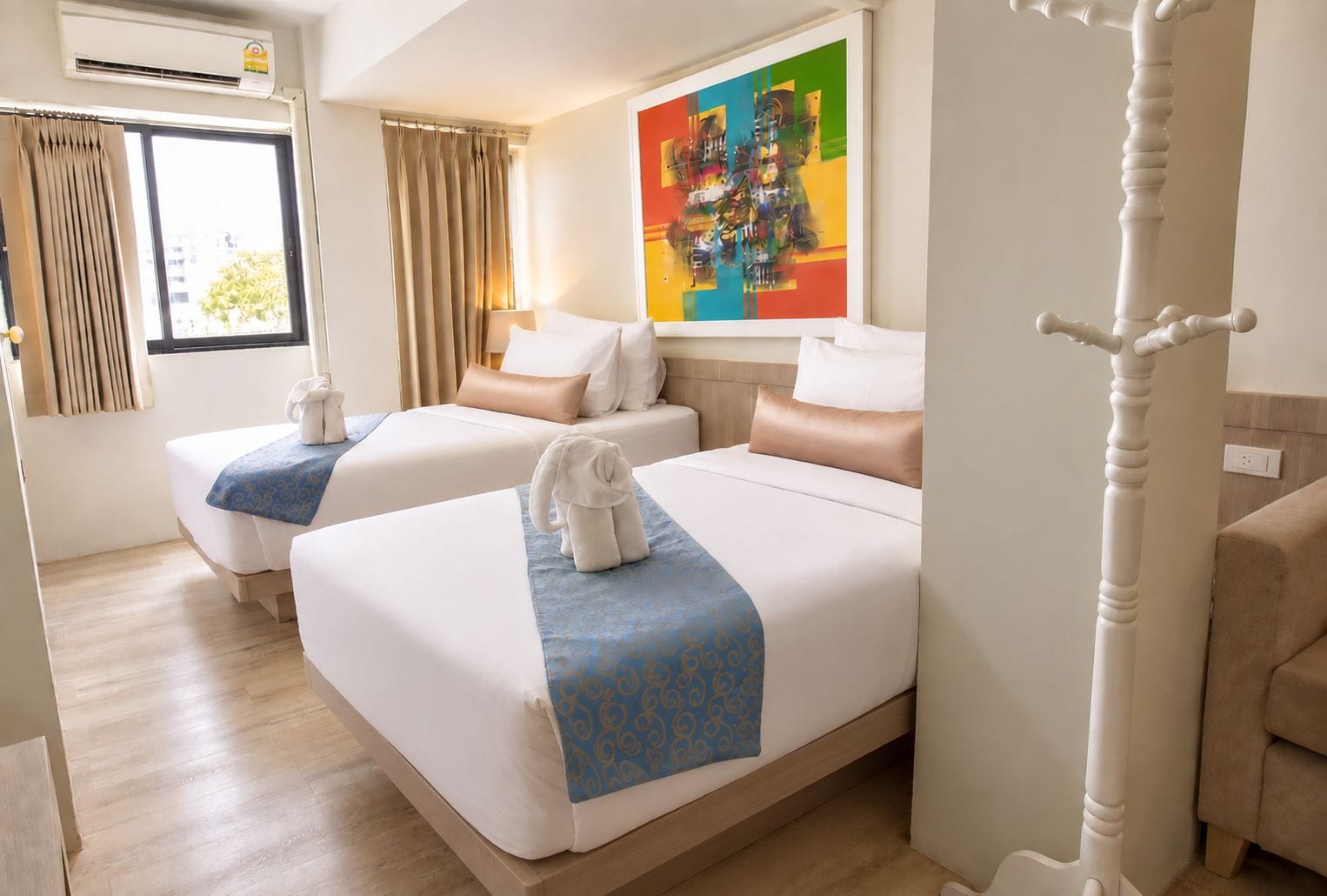 A hotel room with two beds, a large window with curtains, a colorful abstract painting on the wall, and a white decorative column.