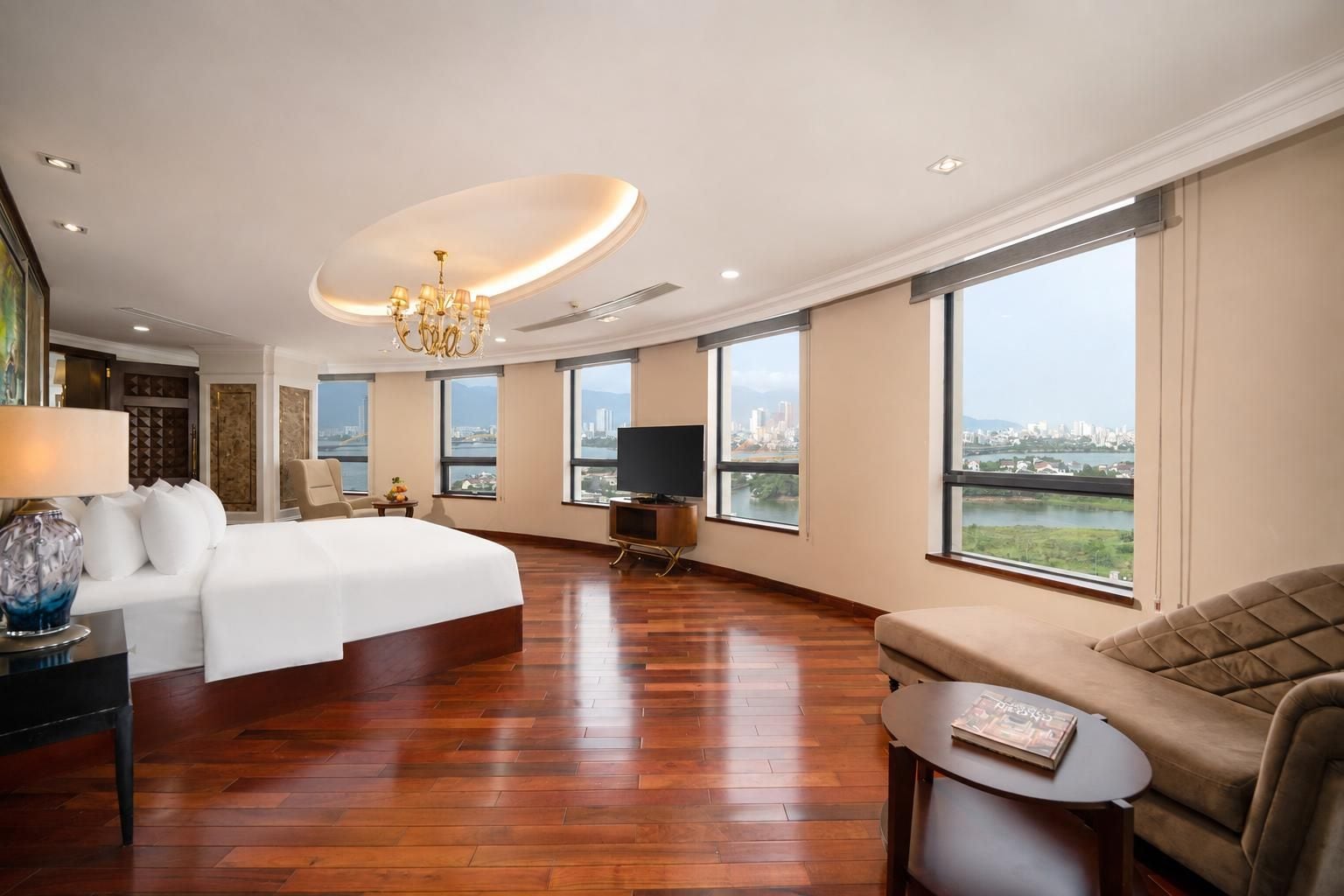 A spacious hotel room with a large bed, a sofa, a television, and a city view.
