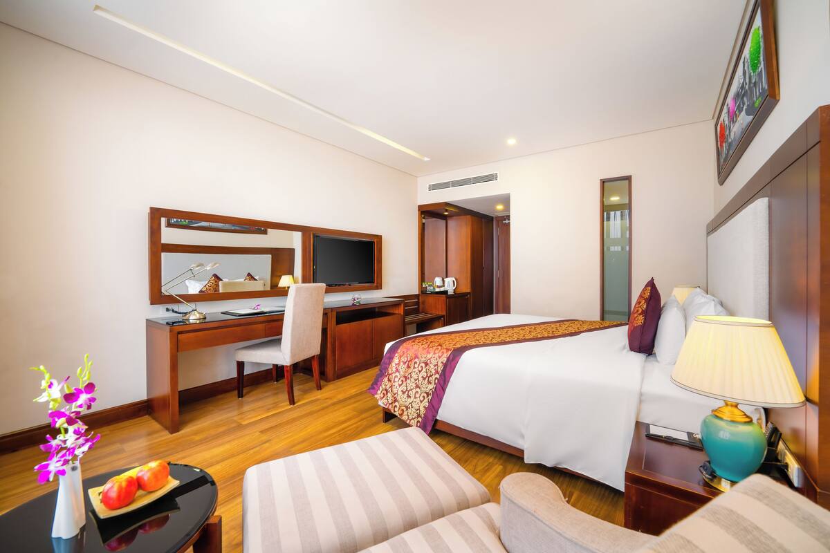 A hotel room with a bed, desk, chair, TV, and a small table with a vase of flowers.
