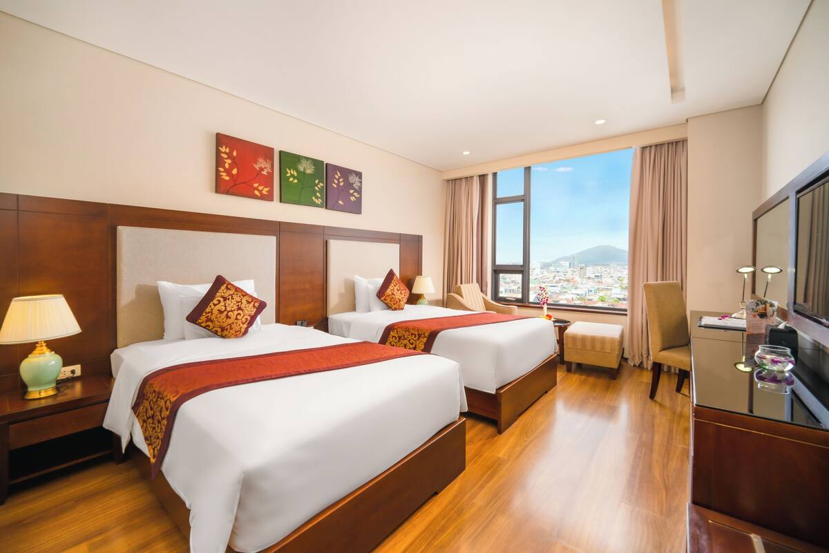 A hotel room with two beds, a desk, a chair, a TV, and a window with a city view.