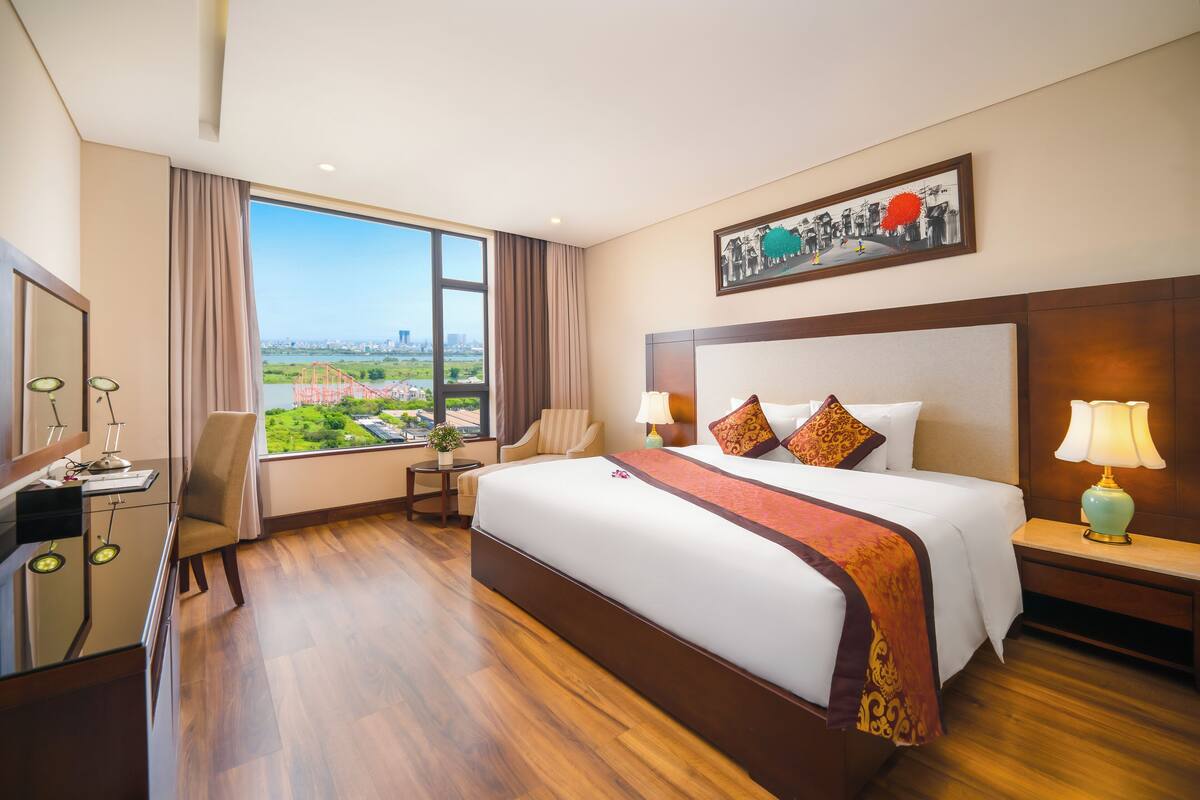 A hotel room with a large bed, a desk, a chair, a TV, and a window with a city view.