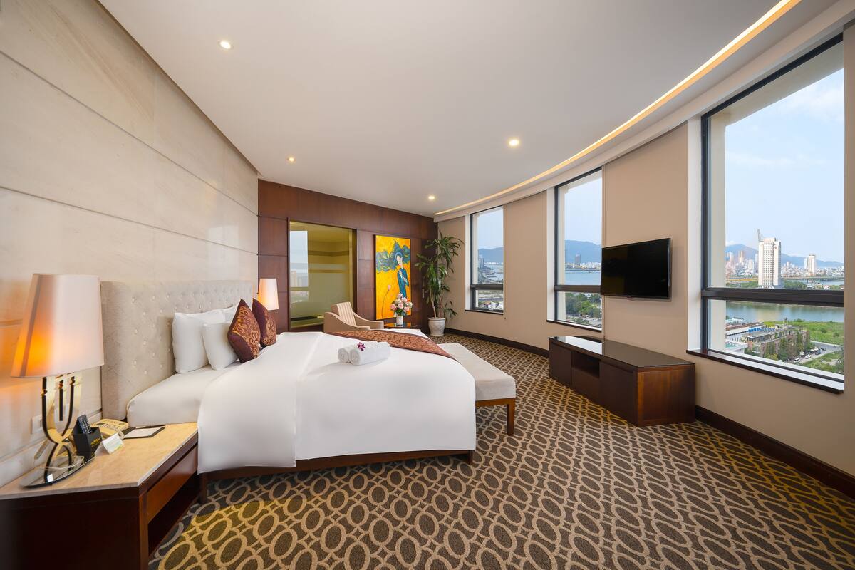 A hotel room with a large bed, a TV, a desk, and a view of the city.