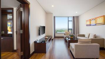 A modern hotel room with a wooden floor, a flat-screen TV, a sofa, a coffee table, and a large window offering a view of the city.