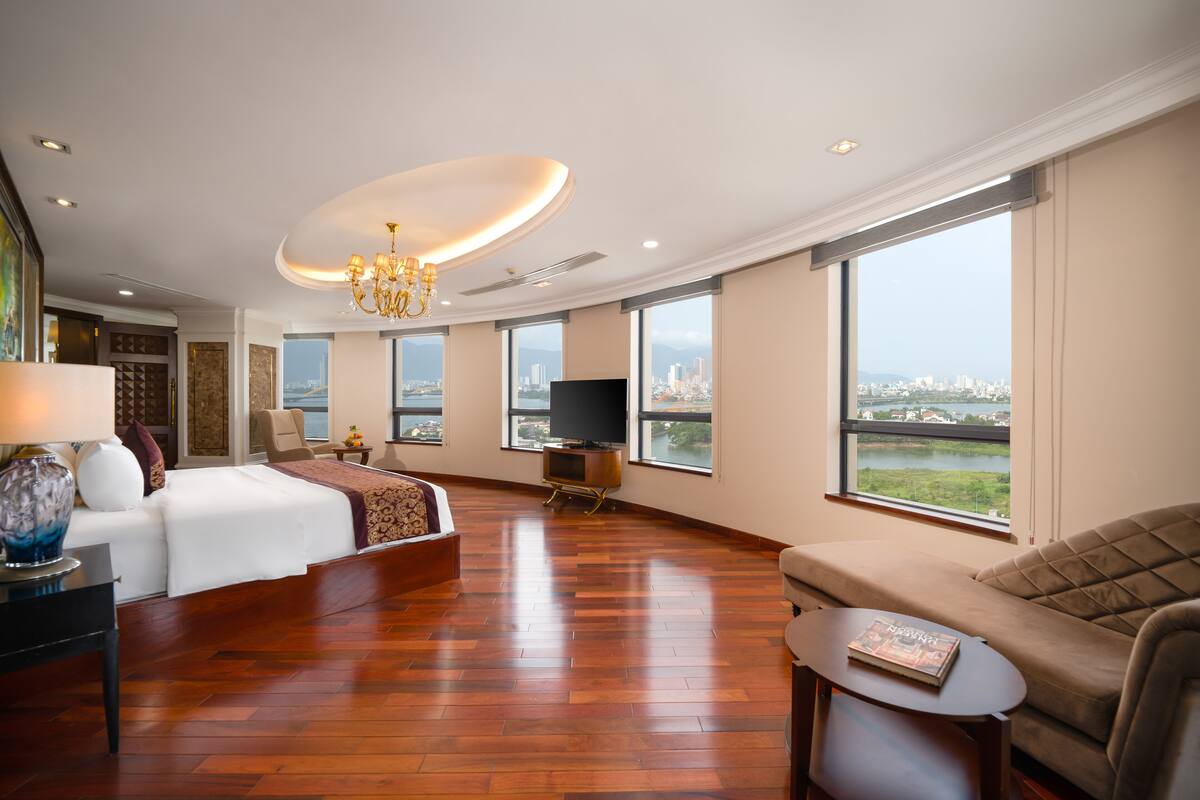 A spacious hotel room with a large bed, a sofa, a TV, and a view of a cityscape through large windows.