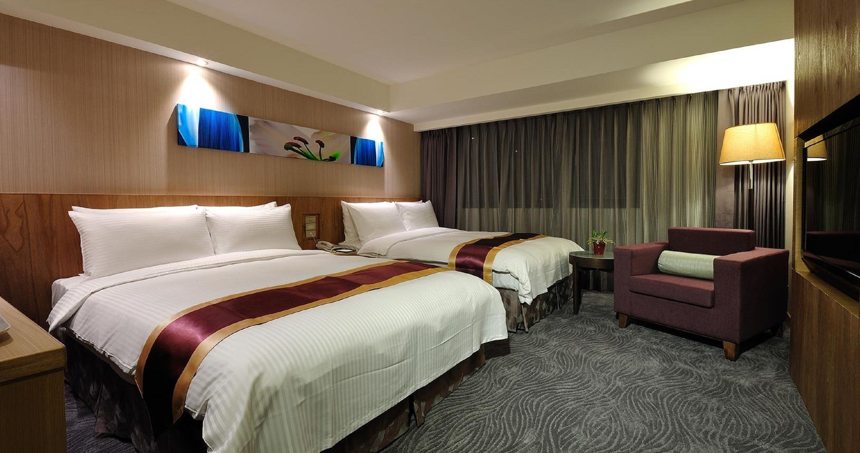 A hotel room with two beds, a small table, a chair, a lamp, and a TV.