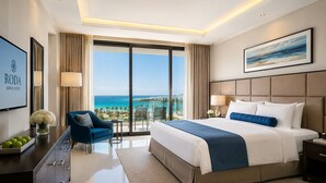 A modern hotel room with a large bed, a TV, a desk with a vase of flowers, and a view of the ocean. - Roda Amwaj Suites (Dubai)