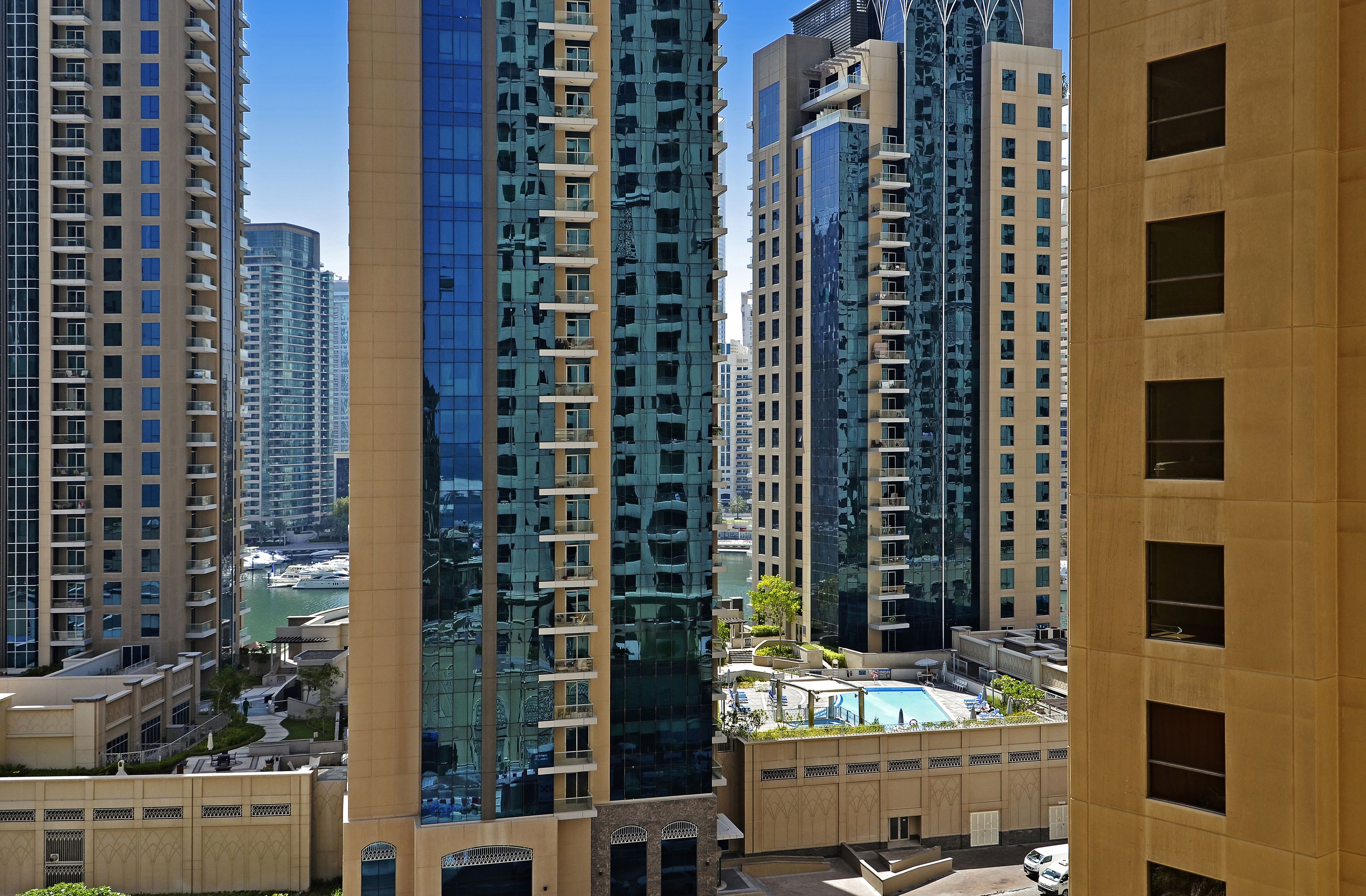 Un edificio residencial alto con varios balcones, una piscina y un puerto deportivo al fondo.