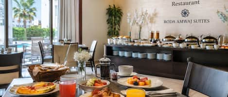Ein Frühstücksbuffet in einem Hotel mit Blick auf Palmen, inklusive verschiedener Speisen. Beschilderung zeigt 'SAMAR Restaurant' und 'RODA AMWAJ SUITES'.
