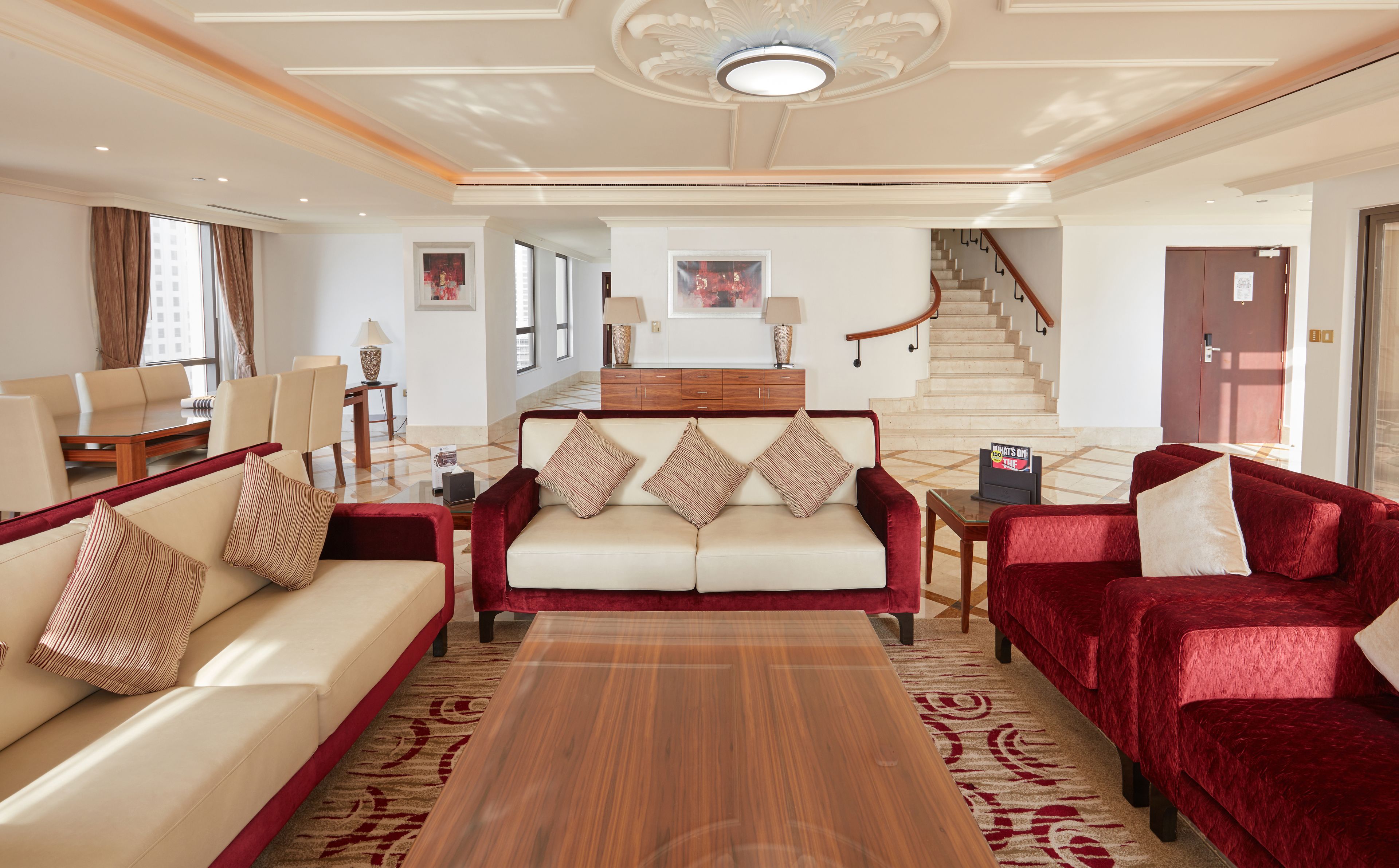 Un vaste espace de vie comprenant un ensemble de canapés, une table basse et un fauteuil en velours rouge. Un meuble en bois, orné d’un tableau encadré, est accroché au-dessus d’un escalier.