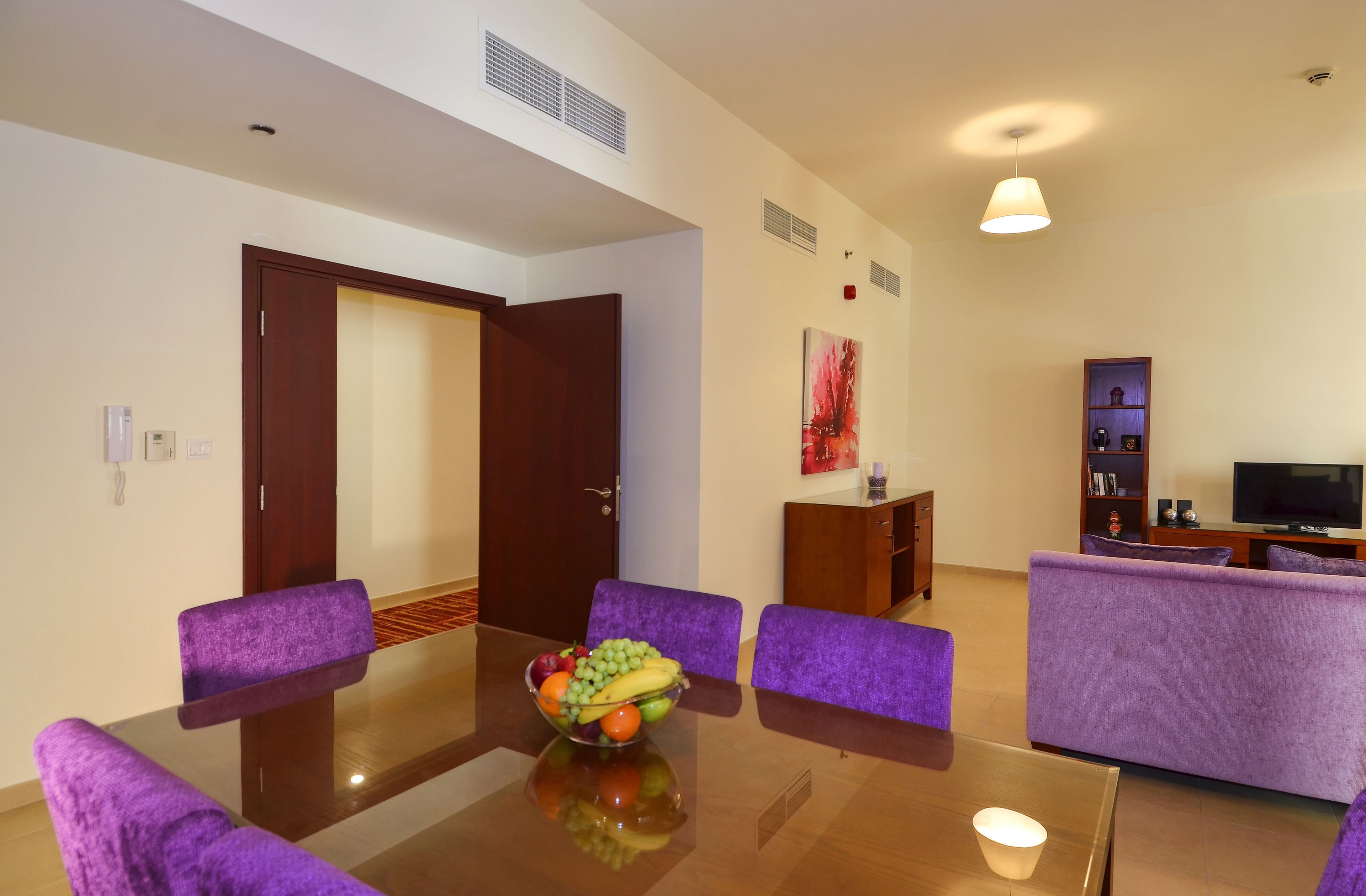A modern living room with a dining area, purple furniture, a fruit bowl on the table, and a television in the background.