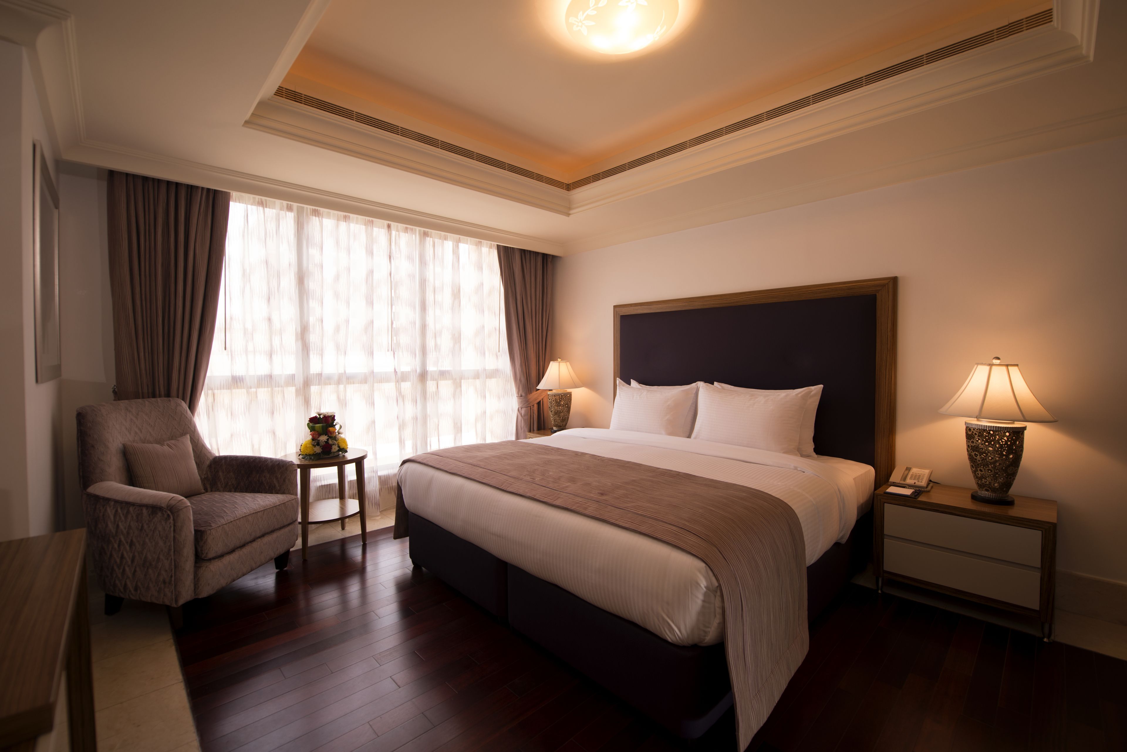 Une chambre d’hôtel avec un grand lit, une chaise, une table de chevet avec une lampe et une fenêtre avec des rideaux.