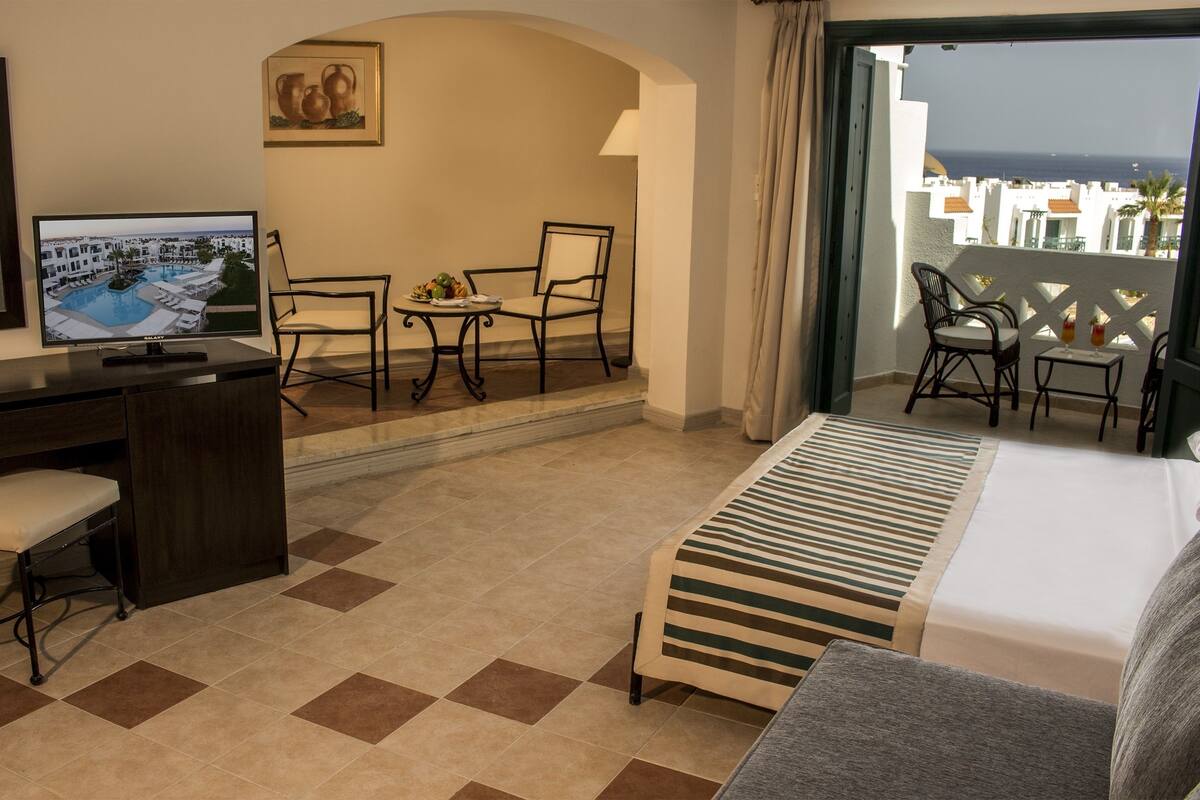 Une chambre d’hôtel équipée d’un lit, d’un bureau, de chaises, d’une télévision et d’un balcon avec vue sur la mer.