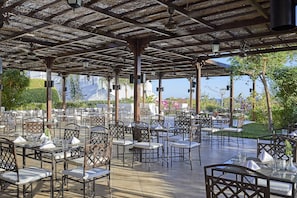 Ein Essbereich im Freien mit Tischen und Stühlen, einer Pergola und Blick auf Grünflächen und Gebäude.