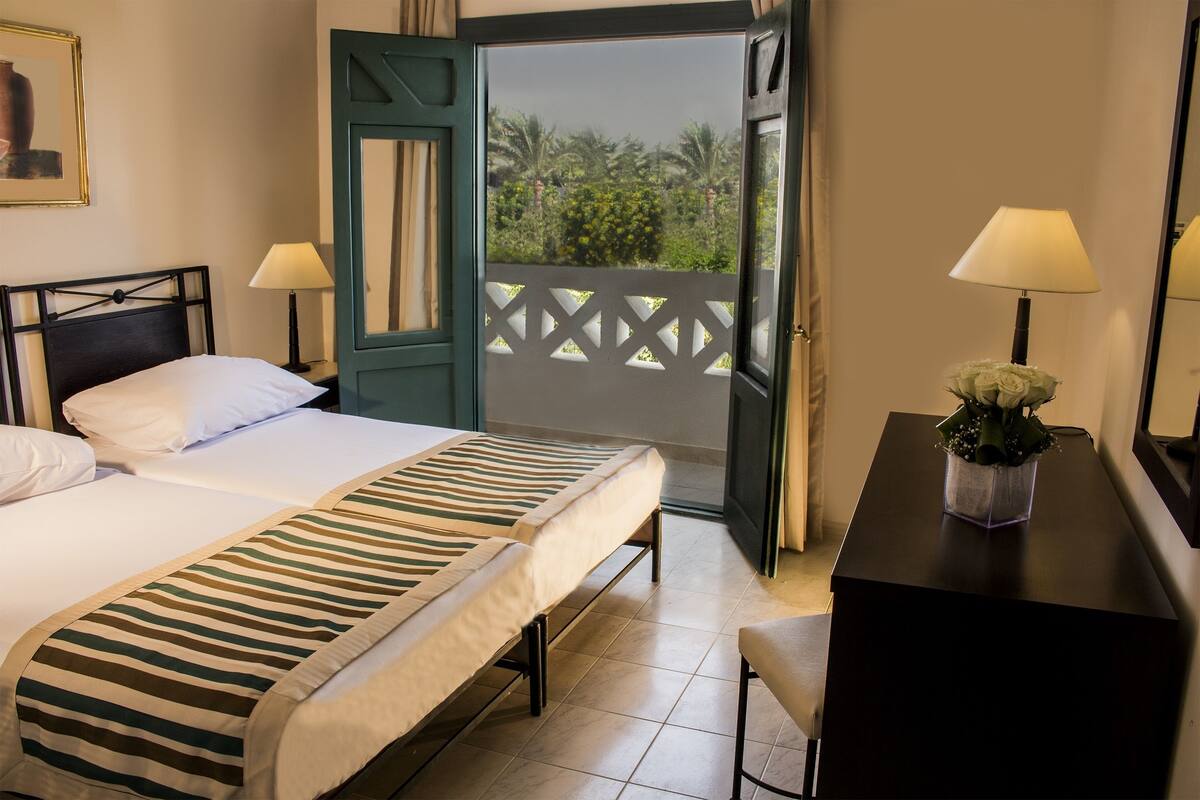 Une chambre d’hôtel avec un lit, des lampes de chevet, un bureau agrémenté d’un vase de fleurs et une vue sur la verdure, visible à travers une porte ouverte.