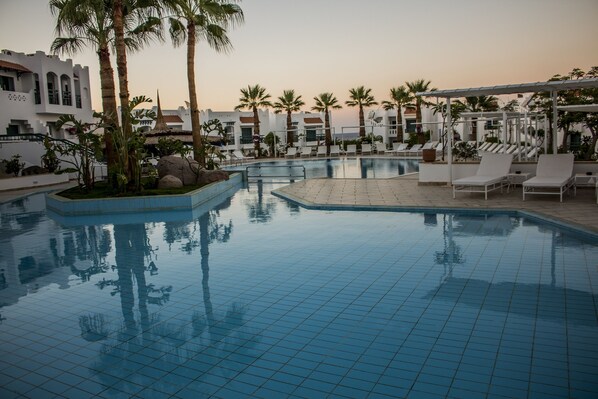 A swimming pool with tiled flooring, palm trees, lounge chairs, and a building with white walls and arched windows.