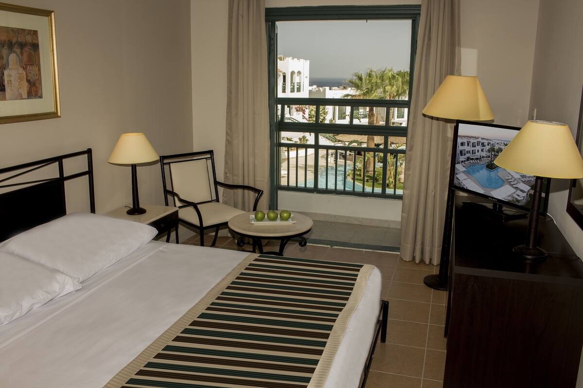 Une chambre d’hôtel avec un lit, deux chaises, une petite table avec des pommes, un bureau avec une lampe et une vue sur une piscine et des immeubles.