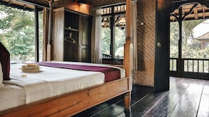 Una cama de madera con colchón blanco y manta roja, una cesta de mimbre con frutas, y vistas al verdor del exterior.