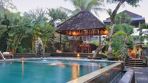 Una piscina con borde de piedra, rodeada de plantas tropicales y una estructura con techo de paja.