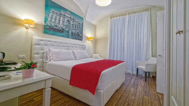 Habitación de hotel con una cama blanca, una manta roja, un sillón blanco, un cuadro en la pared y una lámpara en el techo.
