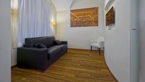 Una sala moderna con un sofá oscuro, una silla blanca, piso de madera, un cuadro grande y una lámpara de techo.