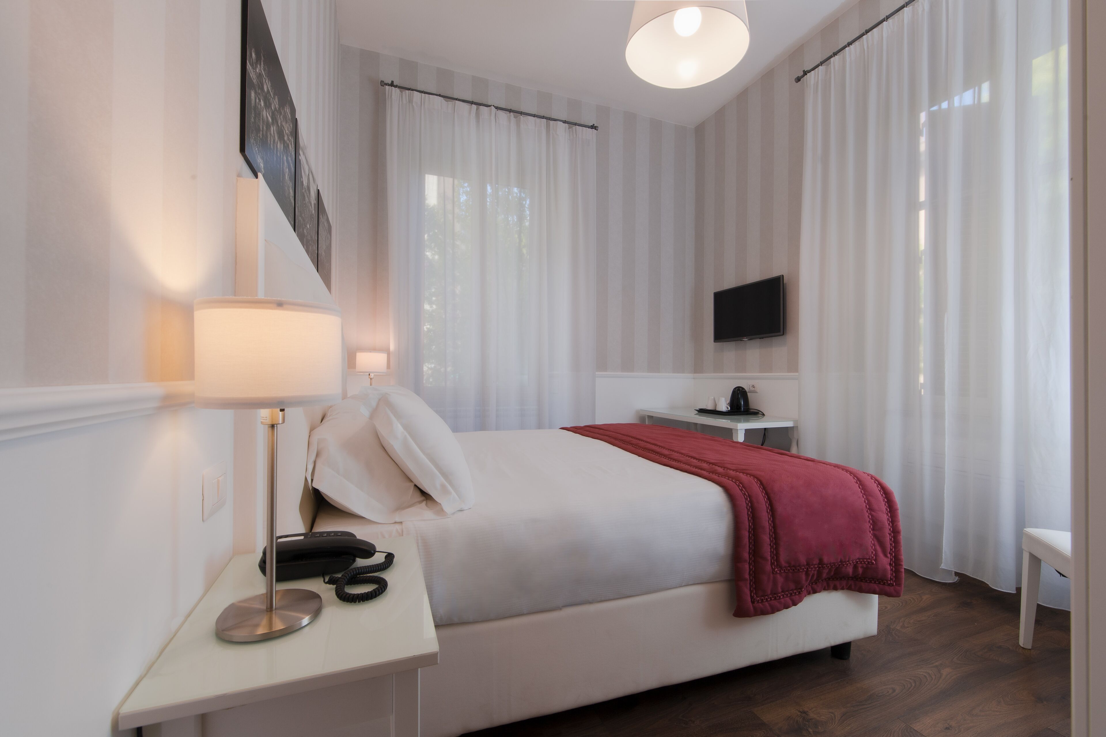 A modern hotel room with a neatly made bed, a bedside table with a lamp and phone, a TV mounted on the wall, and a small desk with a chair.
