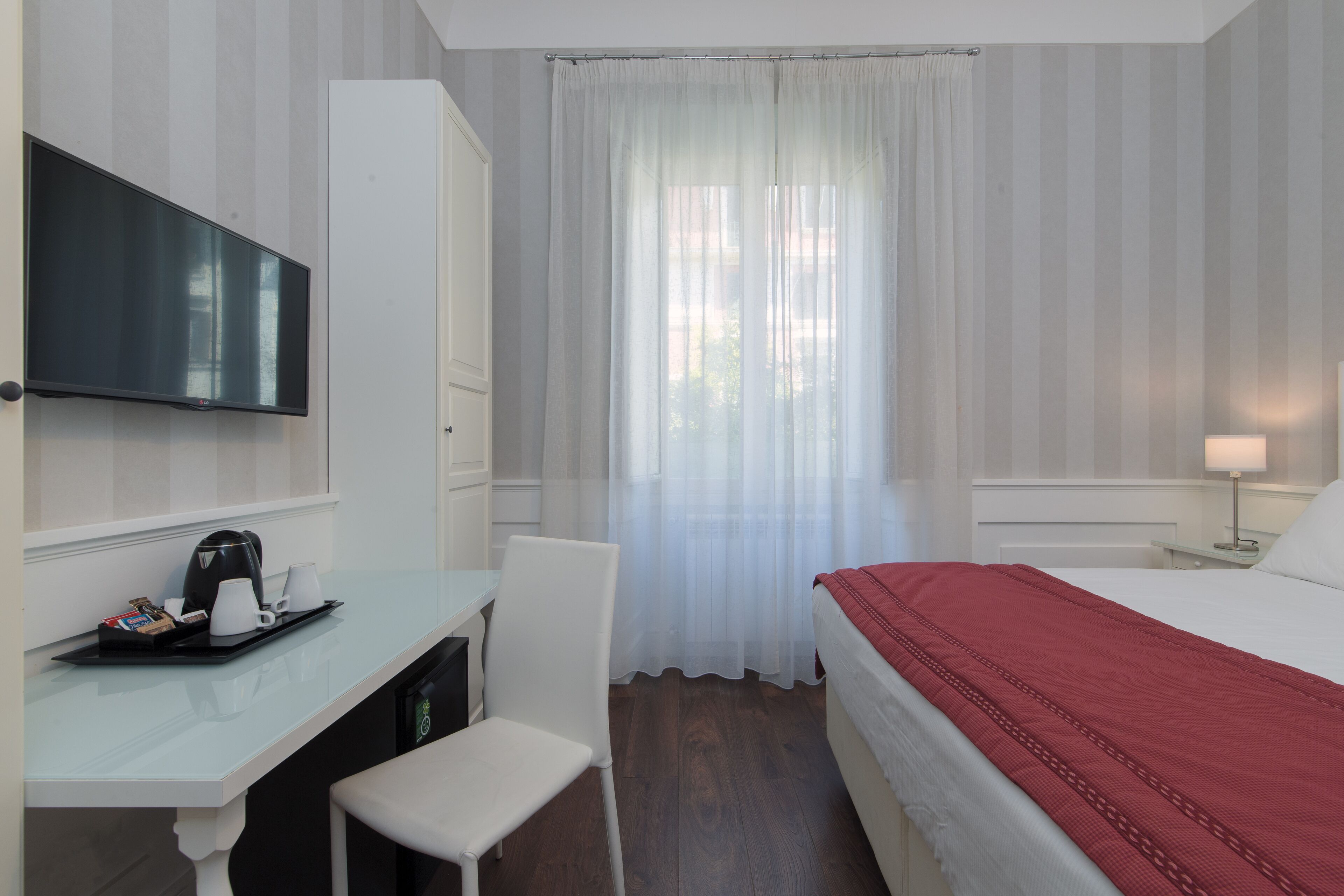 A hotel room with a bed, a desk with a coffee maker, a chair, a TV, and a window with curtains.