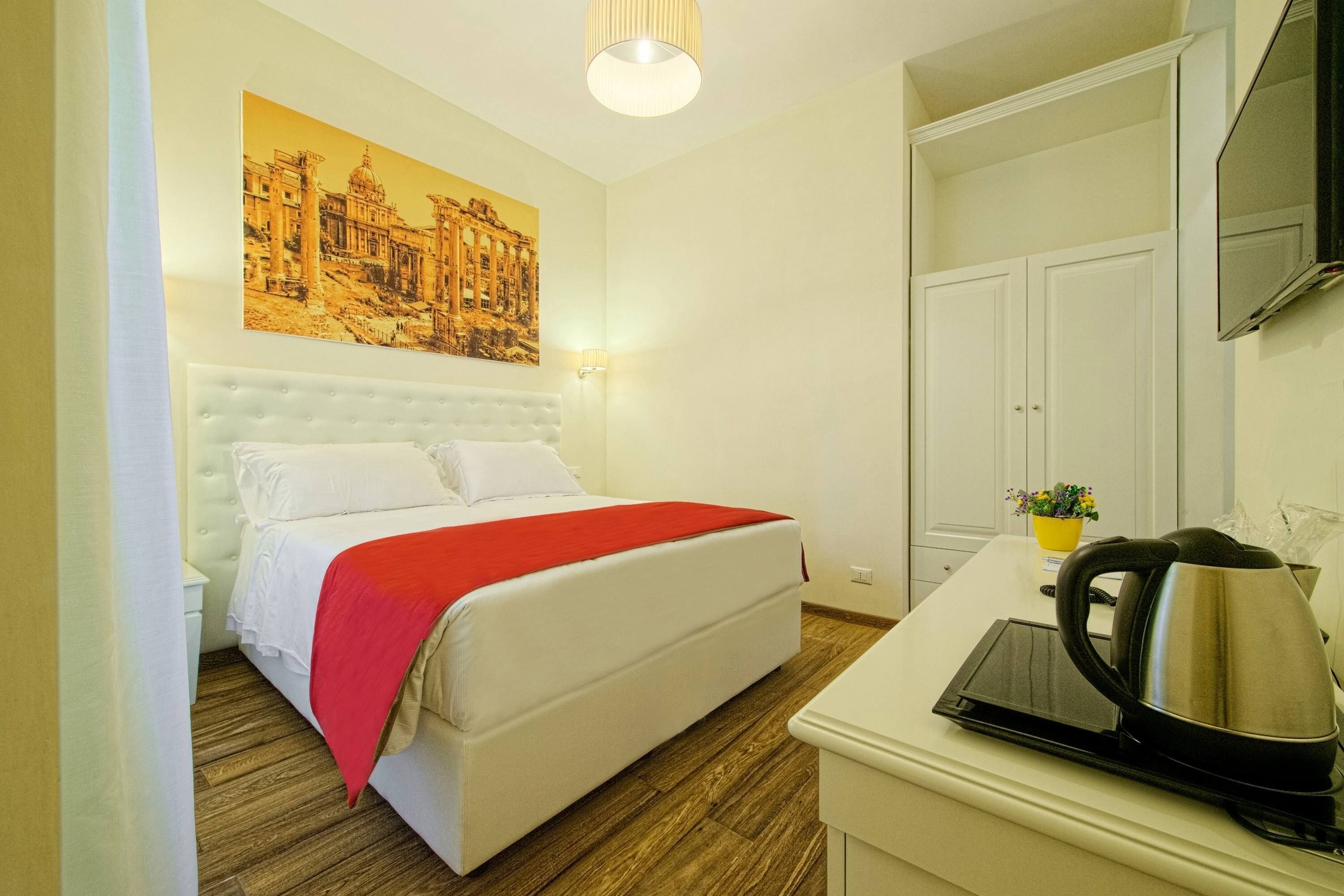 A hotel room with a bed, a bedside table with a kettle, a flat-screen TV, and a painting on the wall.