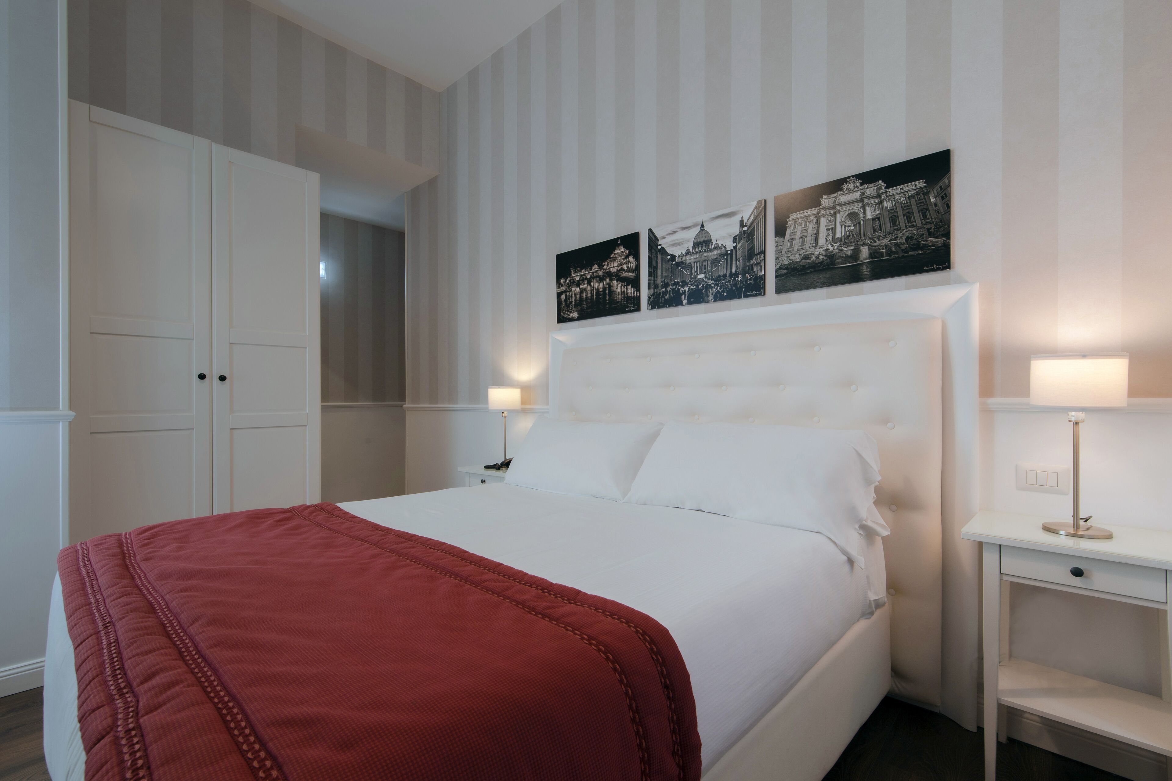 Ein Hotelzimmer mit einem großen Bett, einem Nachttisch mit Lampe und einem Kleiderschrank. Über dem Bett hängen drei schwarz-weiße Fotografien an der Wand.