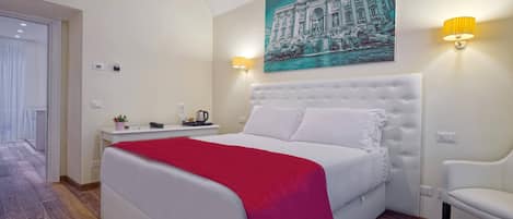 Une chambre d’hôtel avec un grand lit, une tête de lit blanche, un plaid rouge, une chaise blanche, un tableau représentant un bâtiment et deux lampes fixées au mur.
