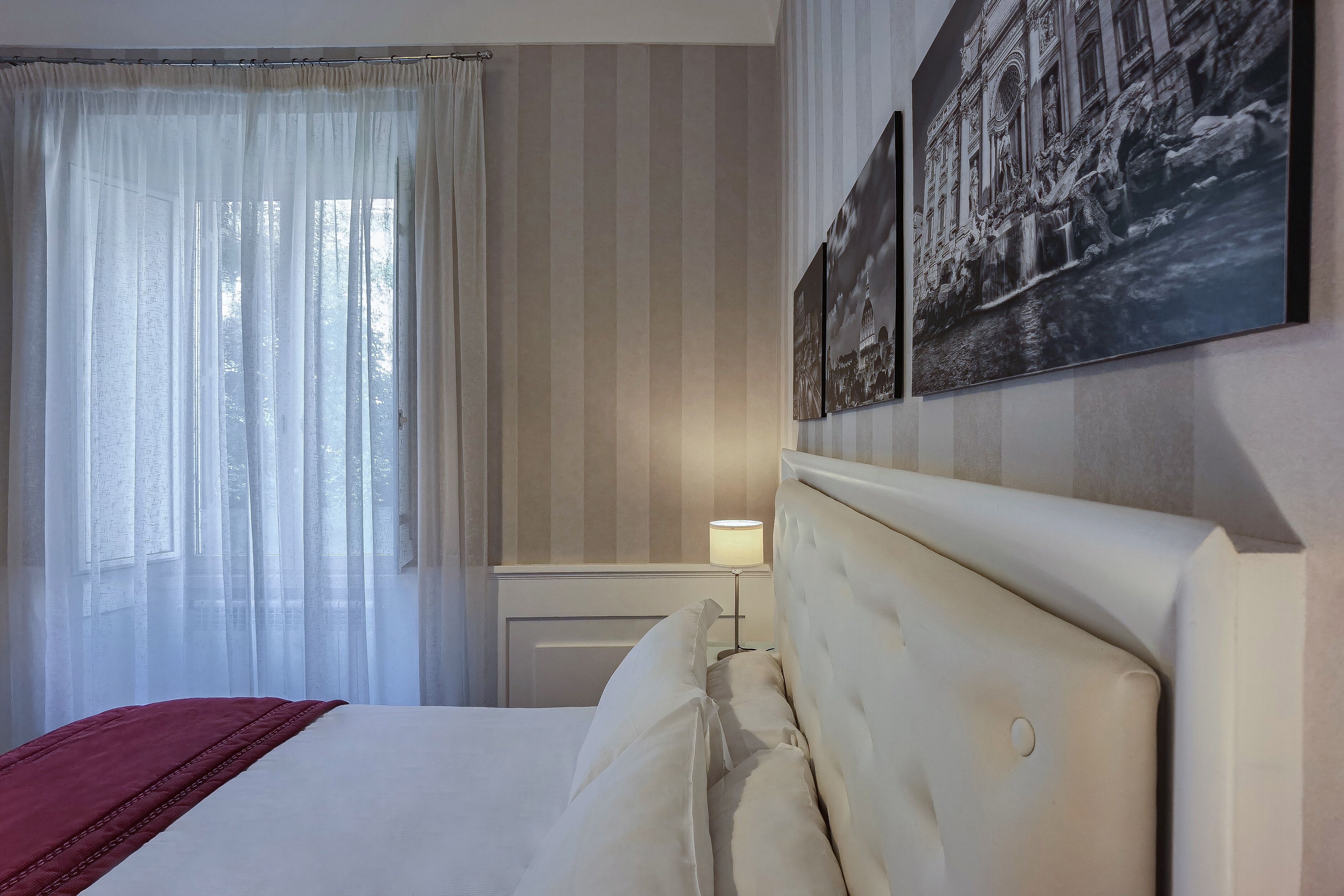 A hotel room with a bed, a window with sheer curtains, a bedside lamp, and two framed pictures on the wall.