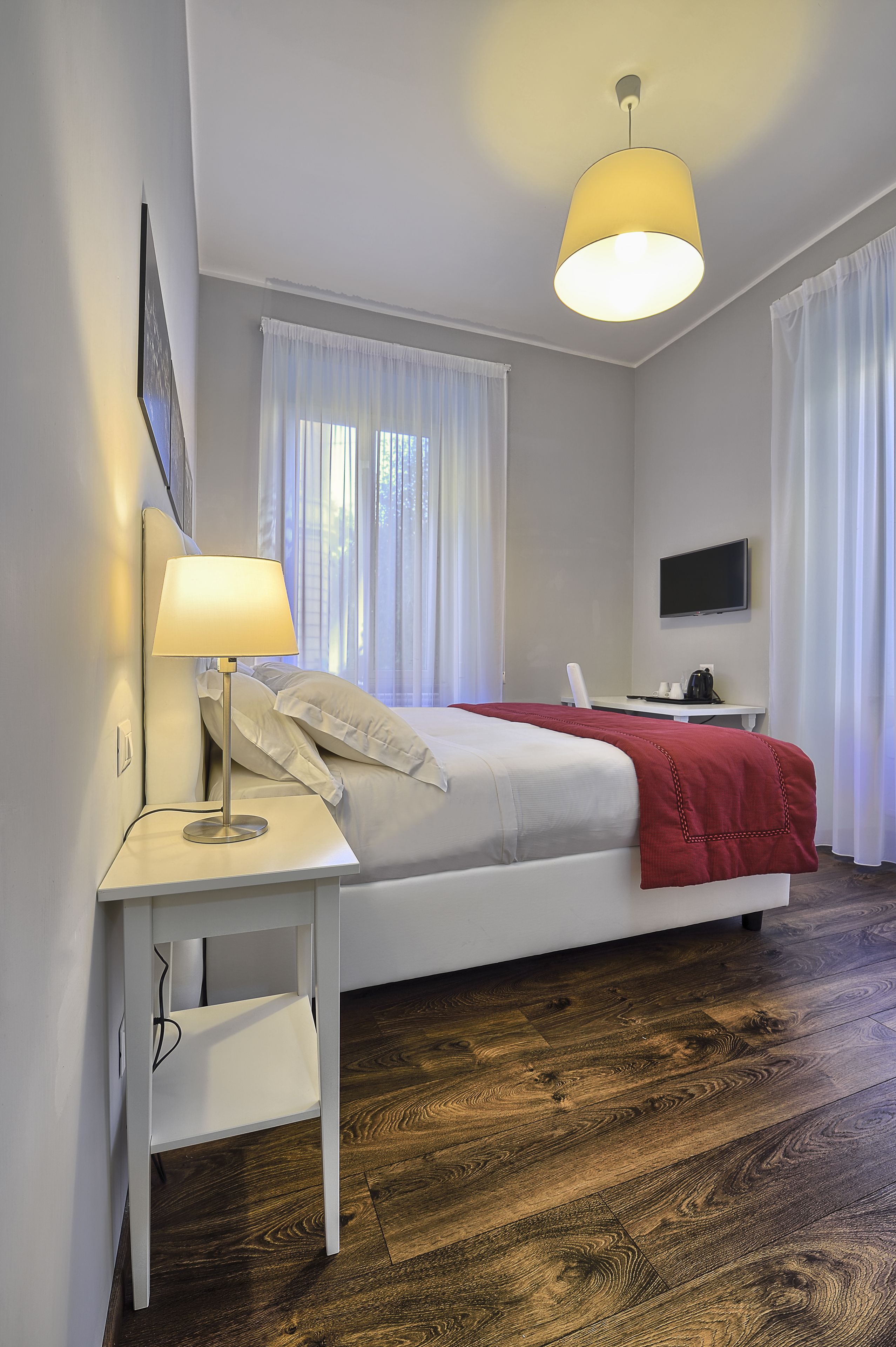 A modern hotel room with a single bed, a desk with a lamp, a TV, and a wooden floor.