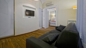 Una habitación moderna con un sofá, un televisor en la pared y una puerta que da a un dormitorio con una cama y una mesita de noche.