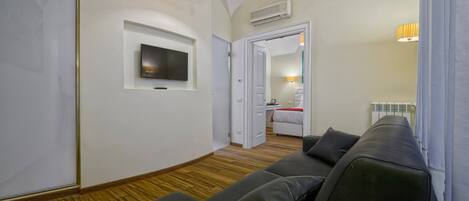 A modern room with a sofa, a TV mounted on the wall, and a door leading to a bedroom with a bed and a nightstand.