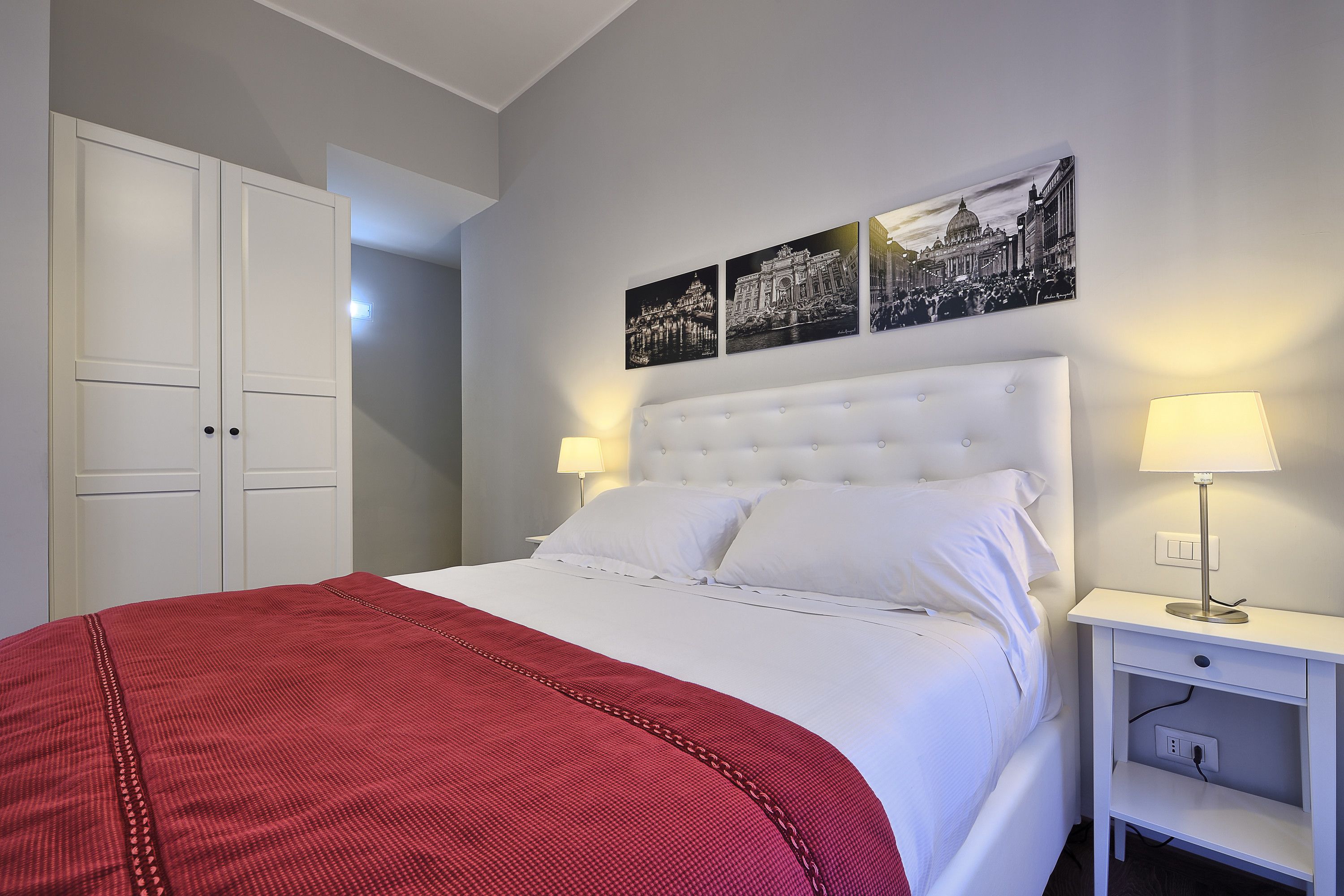 A neatly made bed with white linens and a red blanket, a white nightstand with a lamp, and a wall with three black and white photographs.