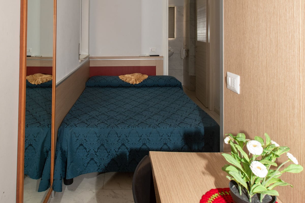 A small, compact bedroom with a bed, a mirror, a wooden desk, and a potted plant.