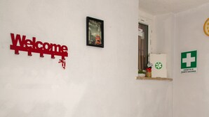 Habitación con escritorio, impresora, cubo de basura, un cartel de bienvenida, un estante pequeño con una planta y un letrero de primeros auxilios.