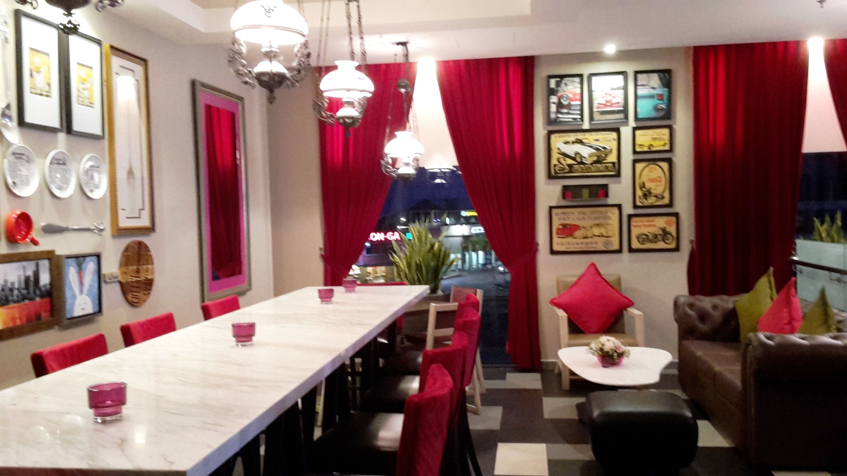 A dining area with a long table, red chairs, and a small white table with a flower. There are framed pictures on the walls and a red curtain in the background.