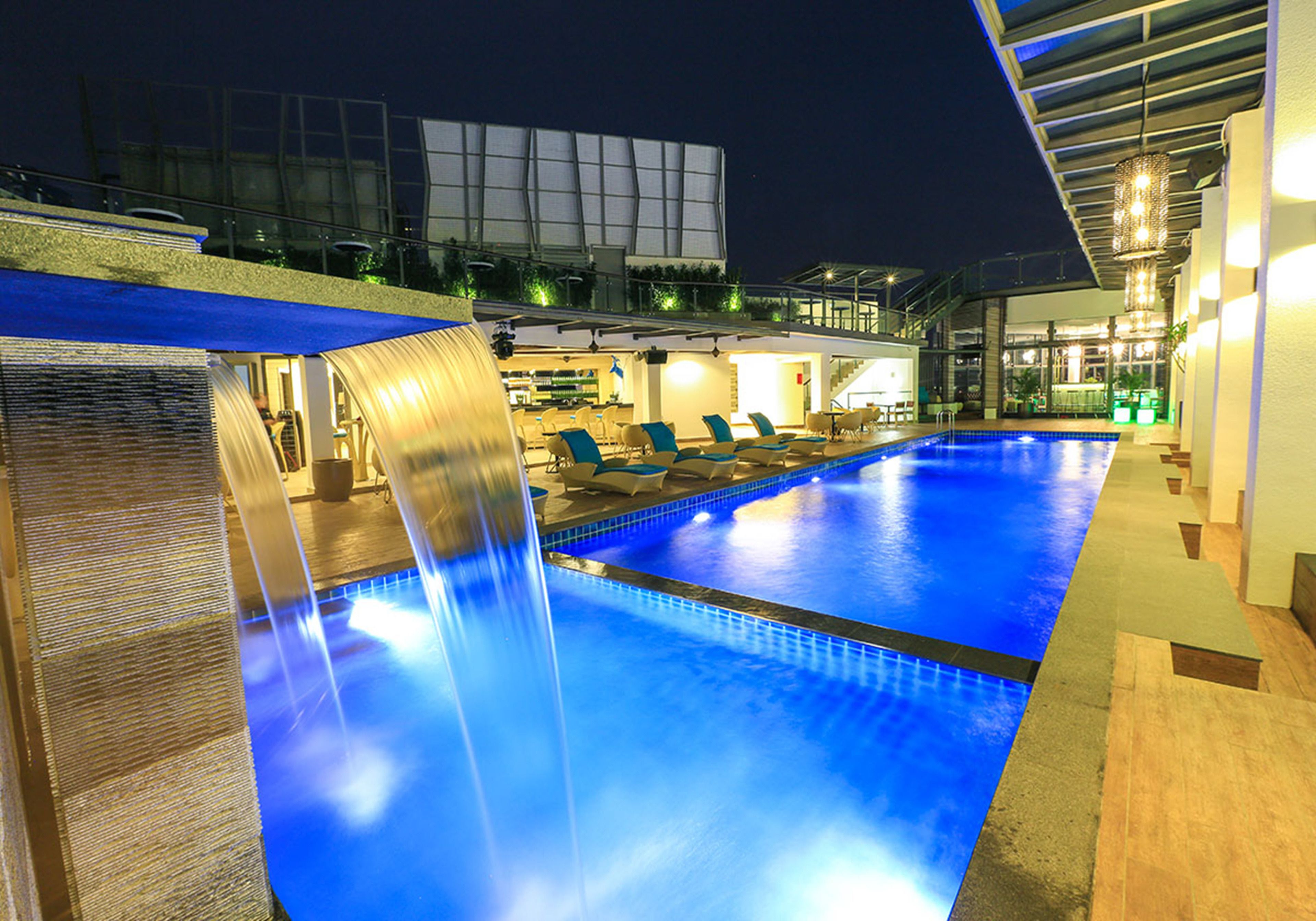 Ein Schwimmbad mit Wasserfall, Liegestühlen und einem modernen Gebäude im Hintergrund.